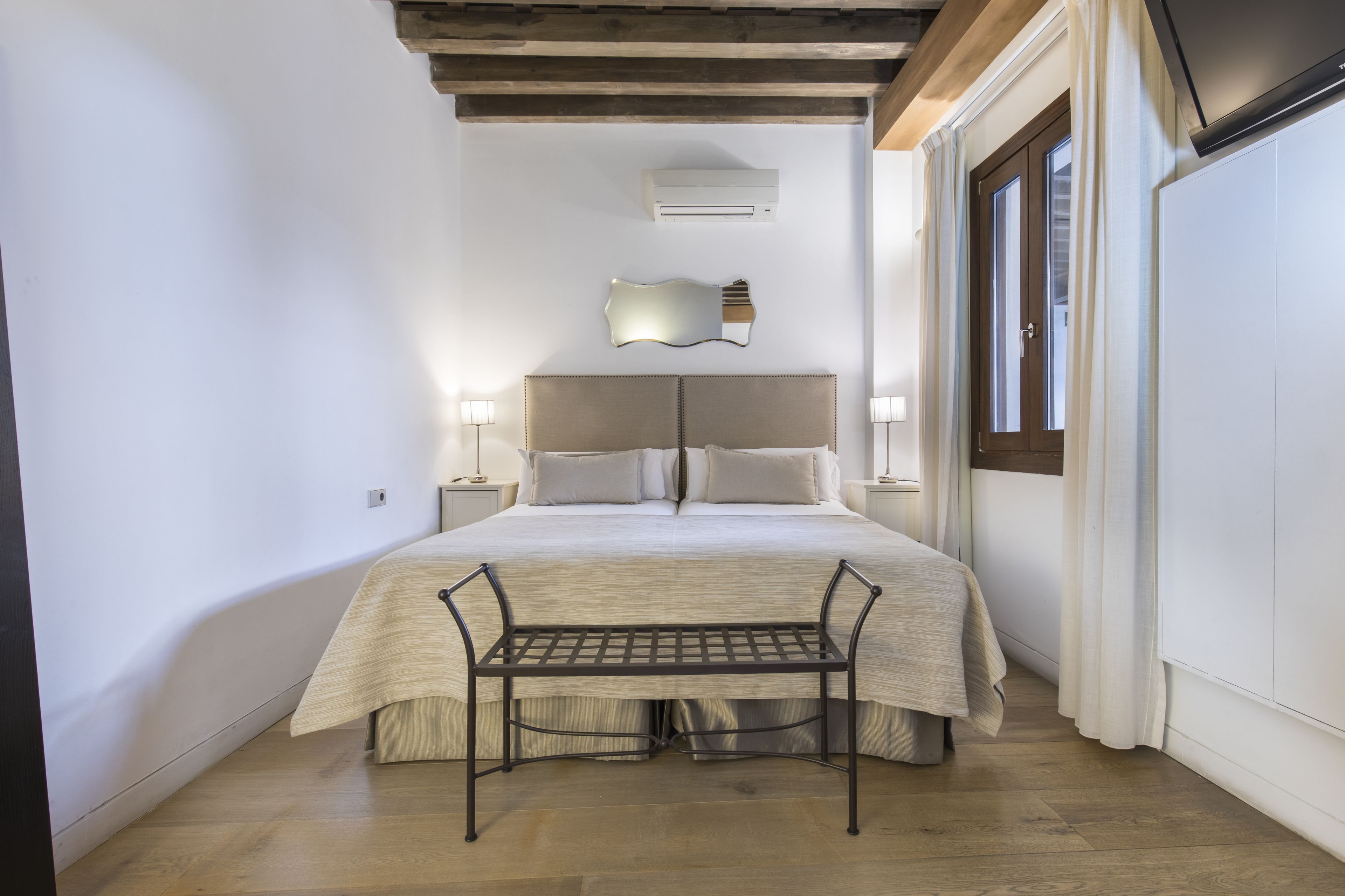 A bedroom with a bed, bedside tables, a bench, and a wall-mounted air conditioner.