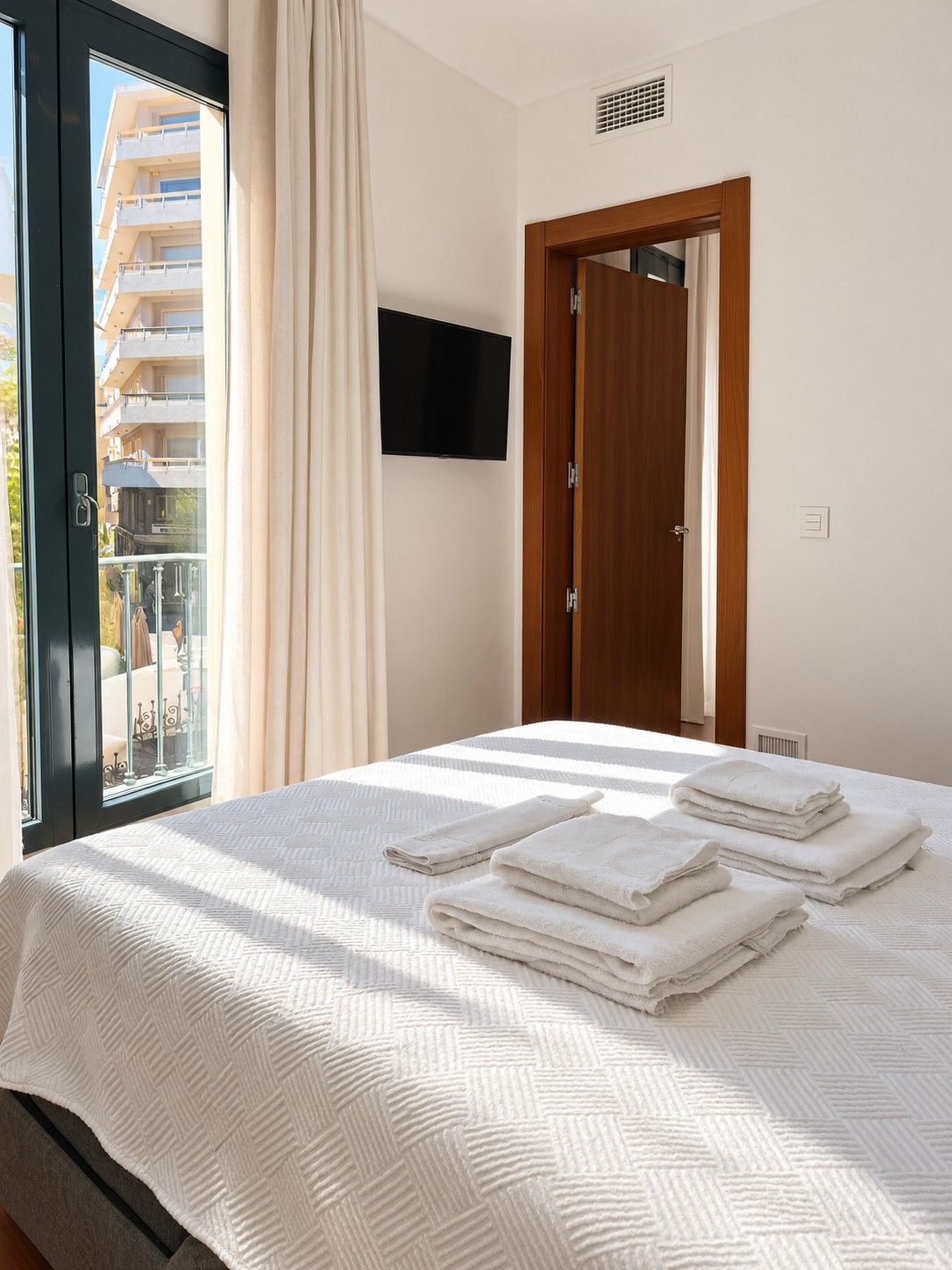 A hotel room with a bed, a television mounted on the wall, a door, and a window with curtains.
