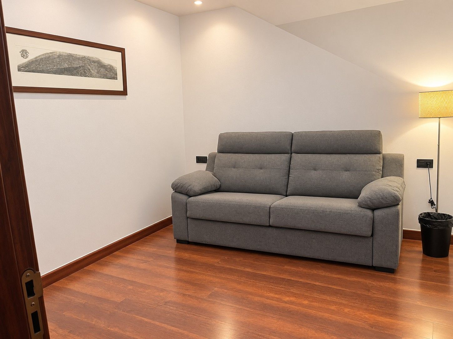 Un canapé aux couleurs neutres dans une pièce dont le sol est recouvert de parquet, avec un tableau encadré au mur et un lampadaire.