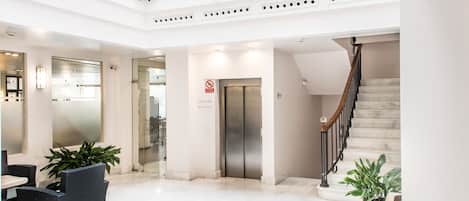 Un moderno vestíbulo de hotel con una escalera de mármol, un mostrador de recepción y un ascensor de vidrio.