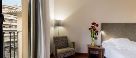 A hotel room with a bed, a chair, a nightstand, a lamp, and a vase with flowers.