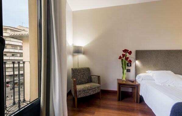 Una habitación de hotel con cama, una silla, una mesita de noche, una lámpara y un florero con flores.