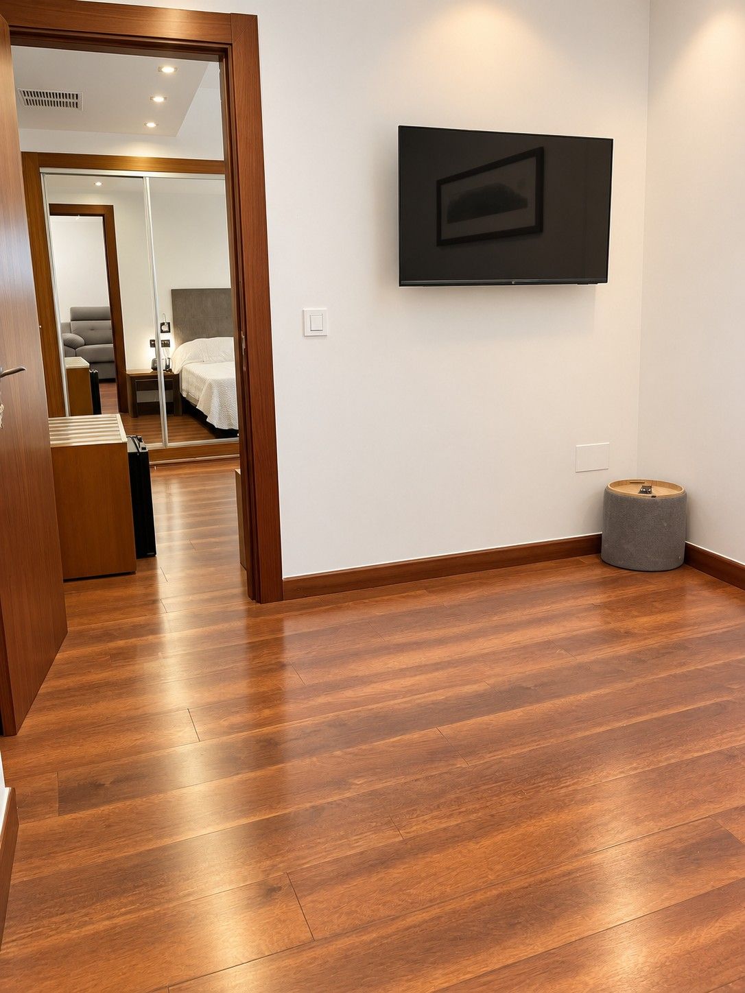 Habitación de hotel con piso de madera, una cama, una mesita de noche, televisor montado en la pared y un taburete pequeño y redondo.