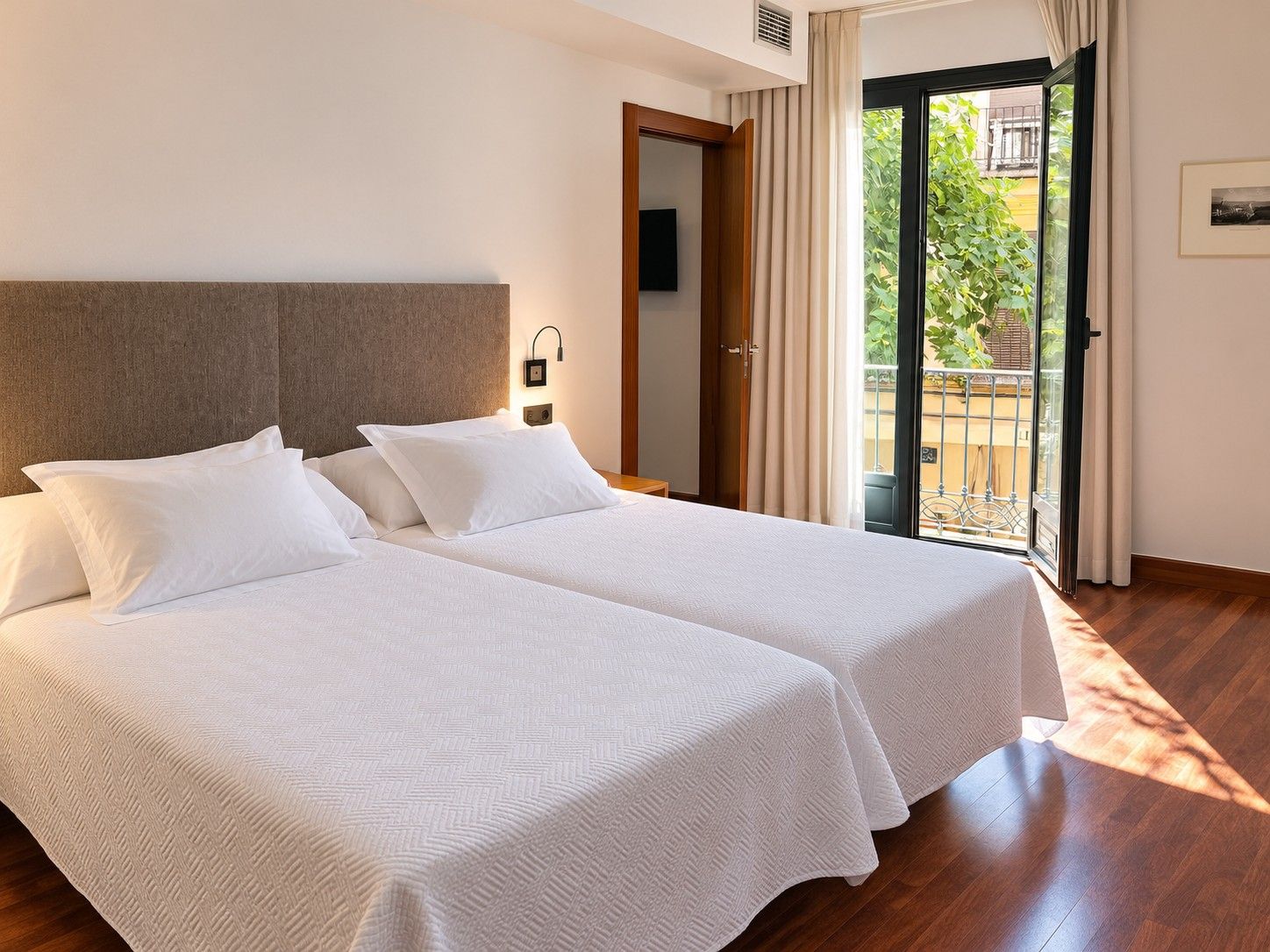 A hotel room with two beds, a wooden headboard, a nightstand, a wall-mounted lamp, and a sliding glass door to a balcony.