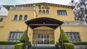 A yellow building with a sign reading "Hotel San Isidro." It features a covered entrance with a metal gate, steps leading up to it, and two trimmed bushes flanking the entrance. - San Isidro Inn (Lima)