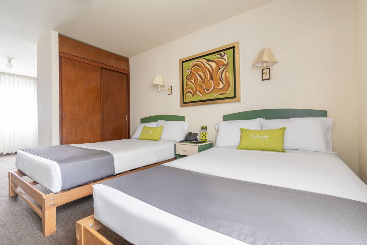 A hotel room with two single beds, a wooden wardrobe, a wall-mounted artwork, and a bedside table with a lamp.