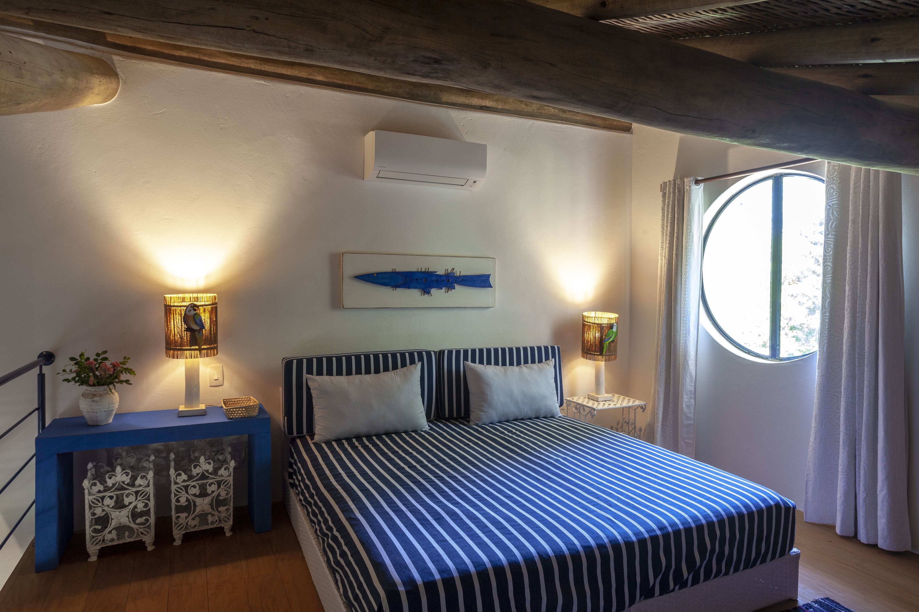 A bedroom with a bed, two bedside tables with lamps, a wall-mounted air conditioner, a window with curtains, and a decorative wall piece.