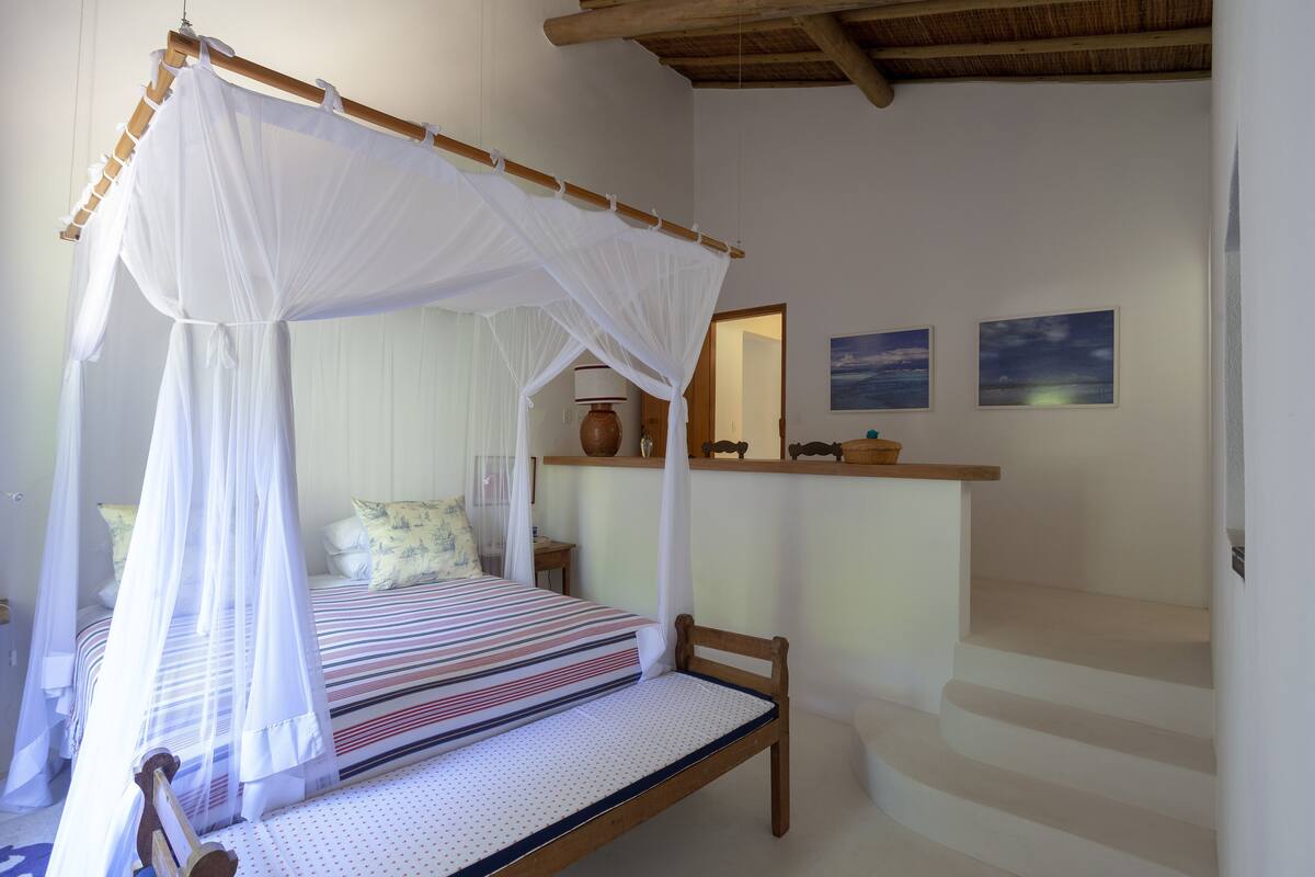 Een slaapkamer met een hemelbed, een houten stoel, een nachtkastje, een plank met decoratieve objecten en een trap.