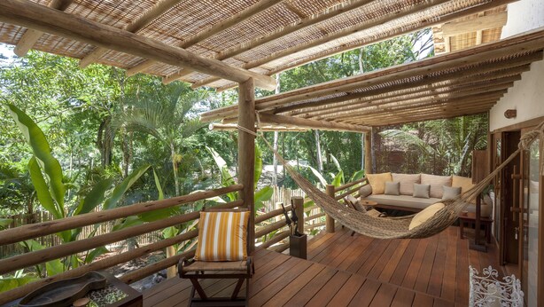 A wooden deck with a hammock, a sofa, and a chair, surrounded by lush greenery. - Etnia Casa Hotel (Porto Seguro)