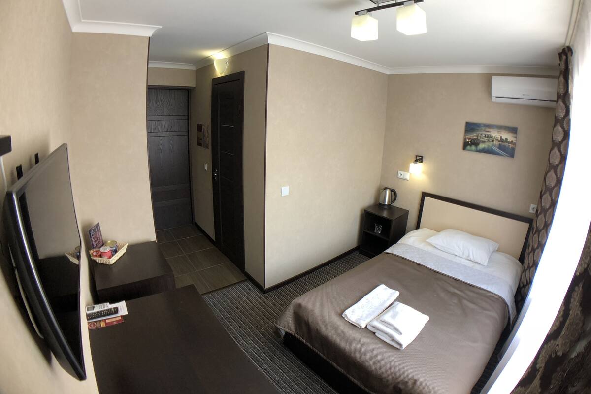 A hotel room with a bed, a desk with a TV, a nightstand with a lamp, and a door leading to another room.