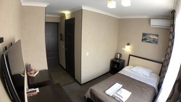 A hotel room with a bed, a desk with a TV, a nightstand with a lamp, and a door leading to another room.