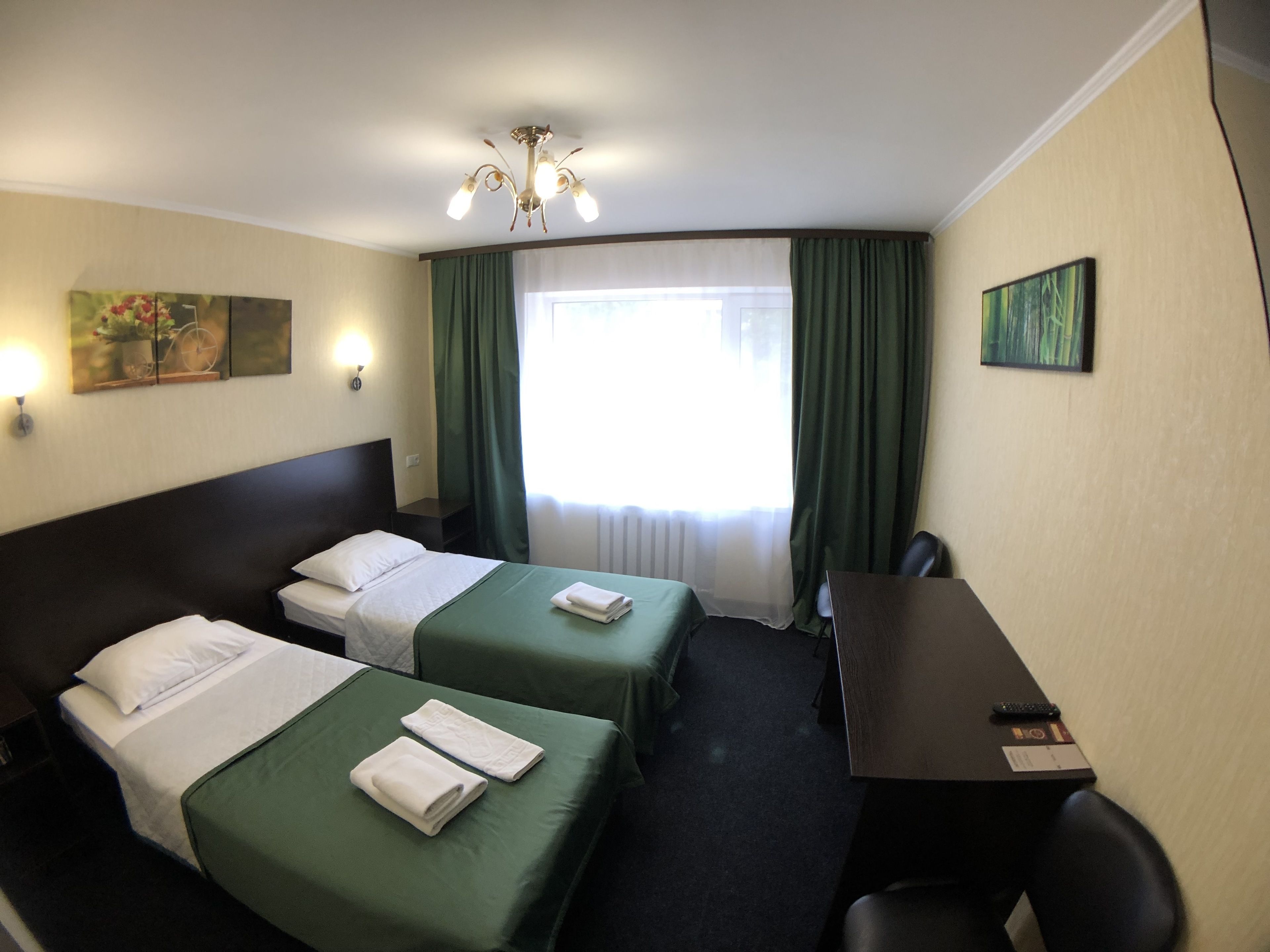 Habitación de hotel con dos camas, un escritorio y una silla. Hay una ventana con cortinas, un cuadro en la pared y una araña.
