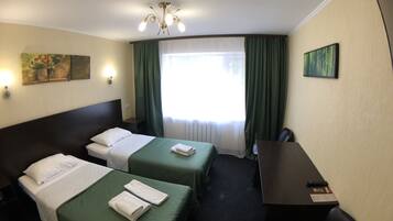 Habitación de hotel con dos camas, un escritorio y una silla. Hay una ventana con cortinas, un cuadro en la pared y una araña.