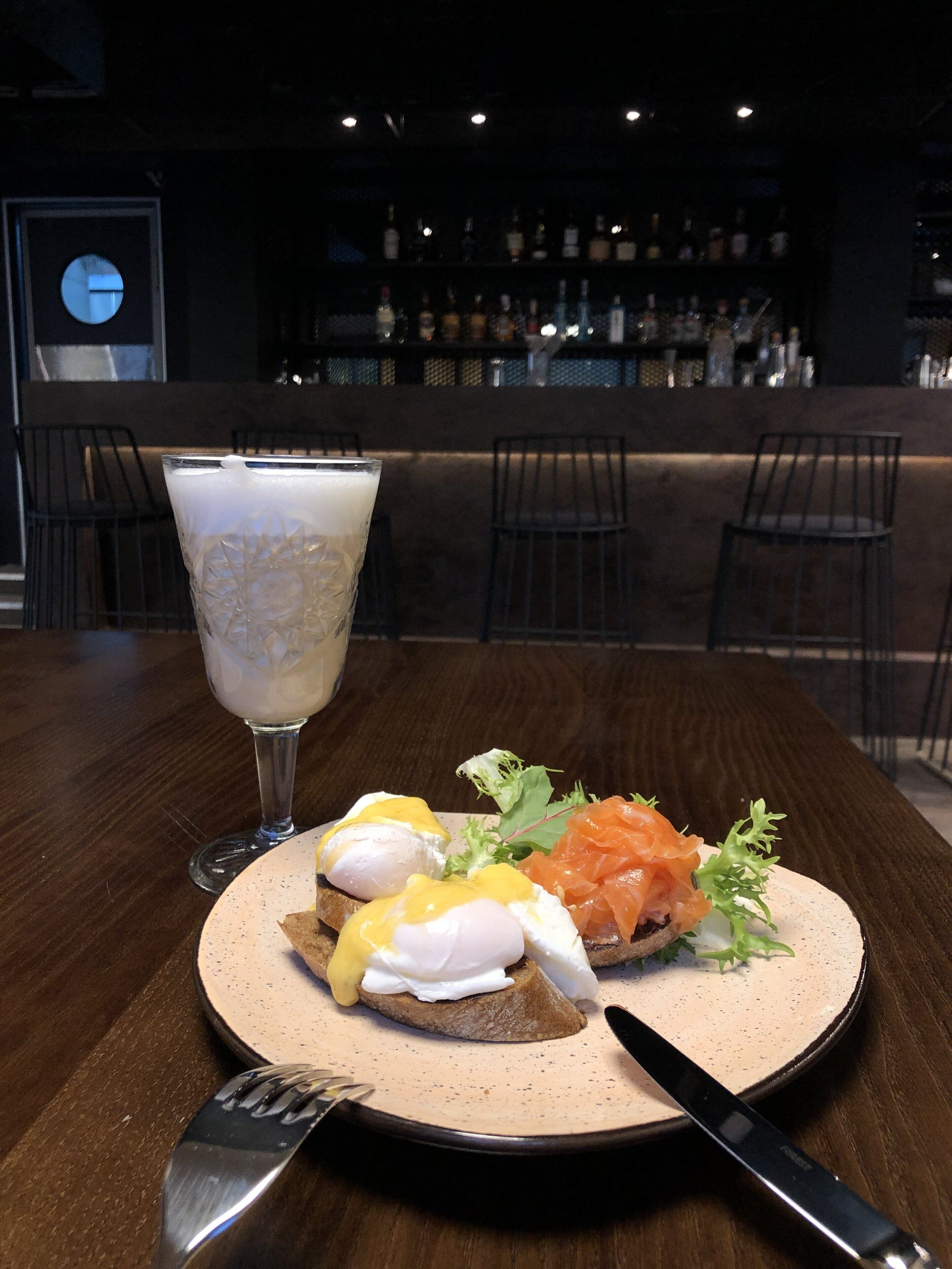 Un desayuno con un plato de huevos Benedict y salmón ahumado, una copa de café helado y una barra de bar al fondo.