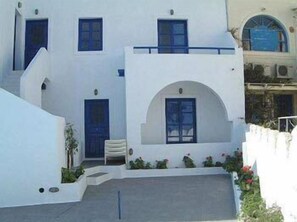 A white building with blue doors and windows, a small balcony, and a paved courtyard. - Olympic Villas (Santorini)