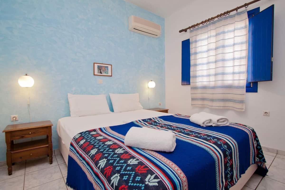 A bedroom with a bed, two bedside tables, a wall-mounted air conditioner, a window with curtains, and a wall-mounted picture.
