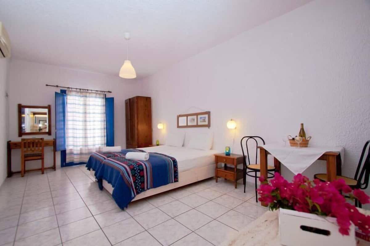 A bedroom with a bed, a desk, a chair, a small table, and a window with curtains.