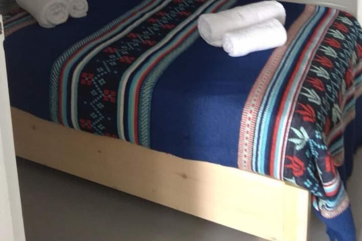 A single bed with a colorful blanket and two rolled towels, a white wall, and a small arched window.