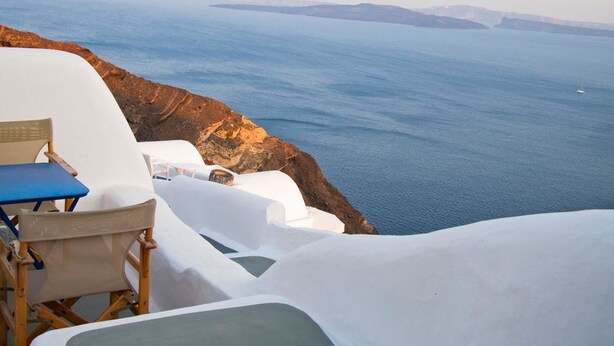 Una estructura blanca y curva con una silla azul y una mesa, con vistas a la costa, acantilados y el mar.
