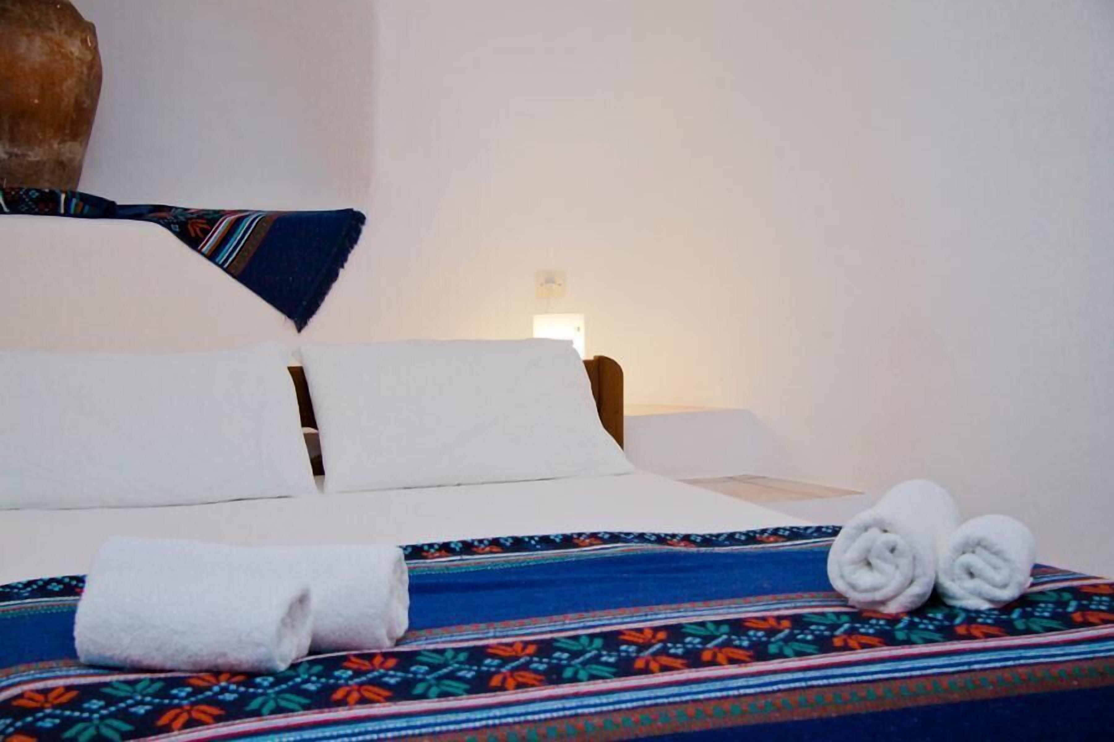 A bed with white linens, a colorful patterned bedspread, and two rolled white towels on the bed.