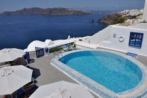 A pool area with a view of the sea and a white building with blue accents. - Olympic Villas (Santorini)