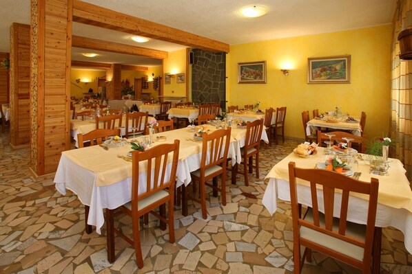 Restaurante com várias mesas postas para refeições, cadeiras de madeira e chão de azulejos de pedra.