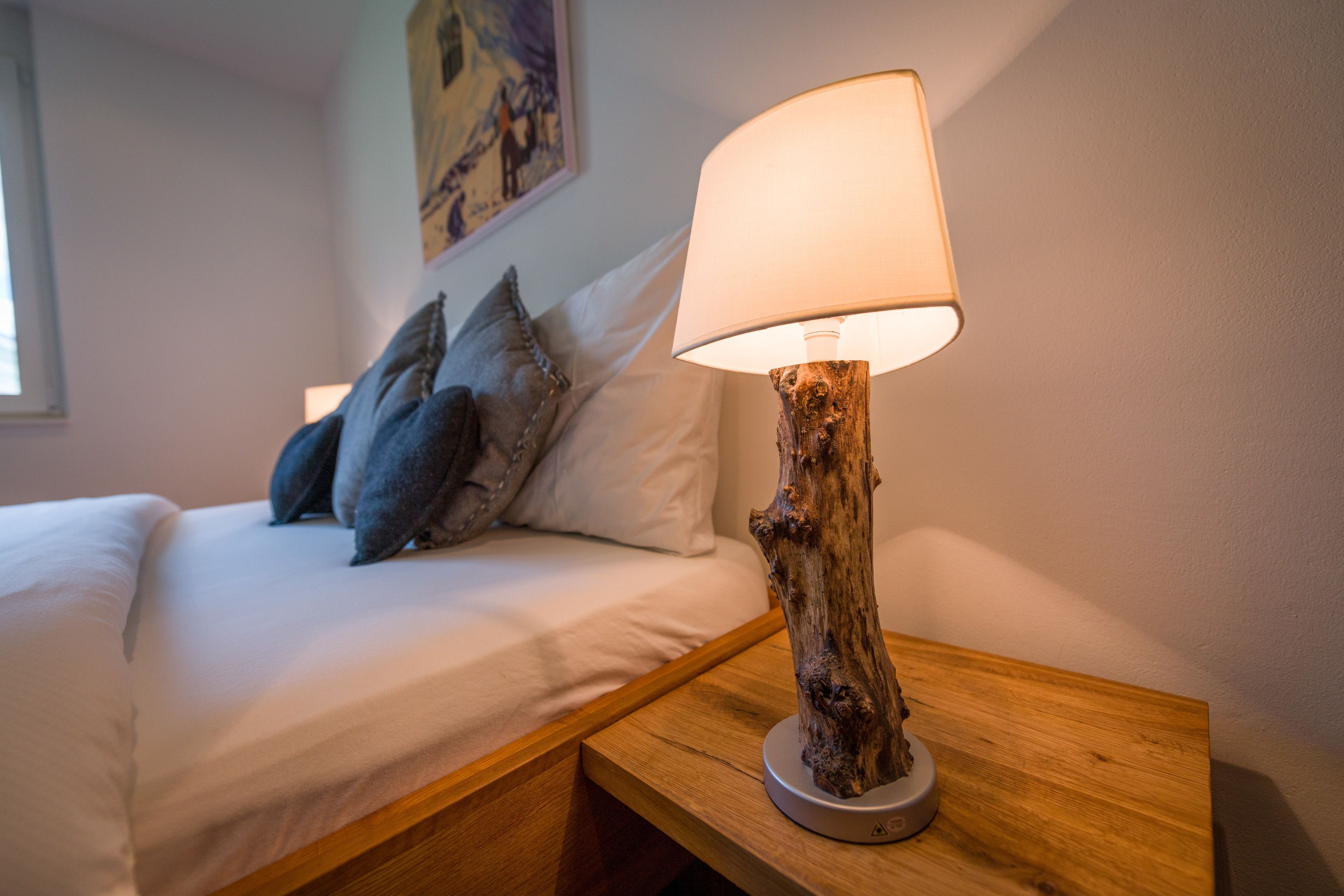 Un lit avec du linge de lit blanc et des oreillers gris, une table de chevet en bois avec une lampe, et un tableau accroché au mur.