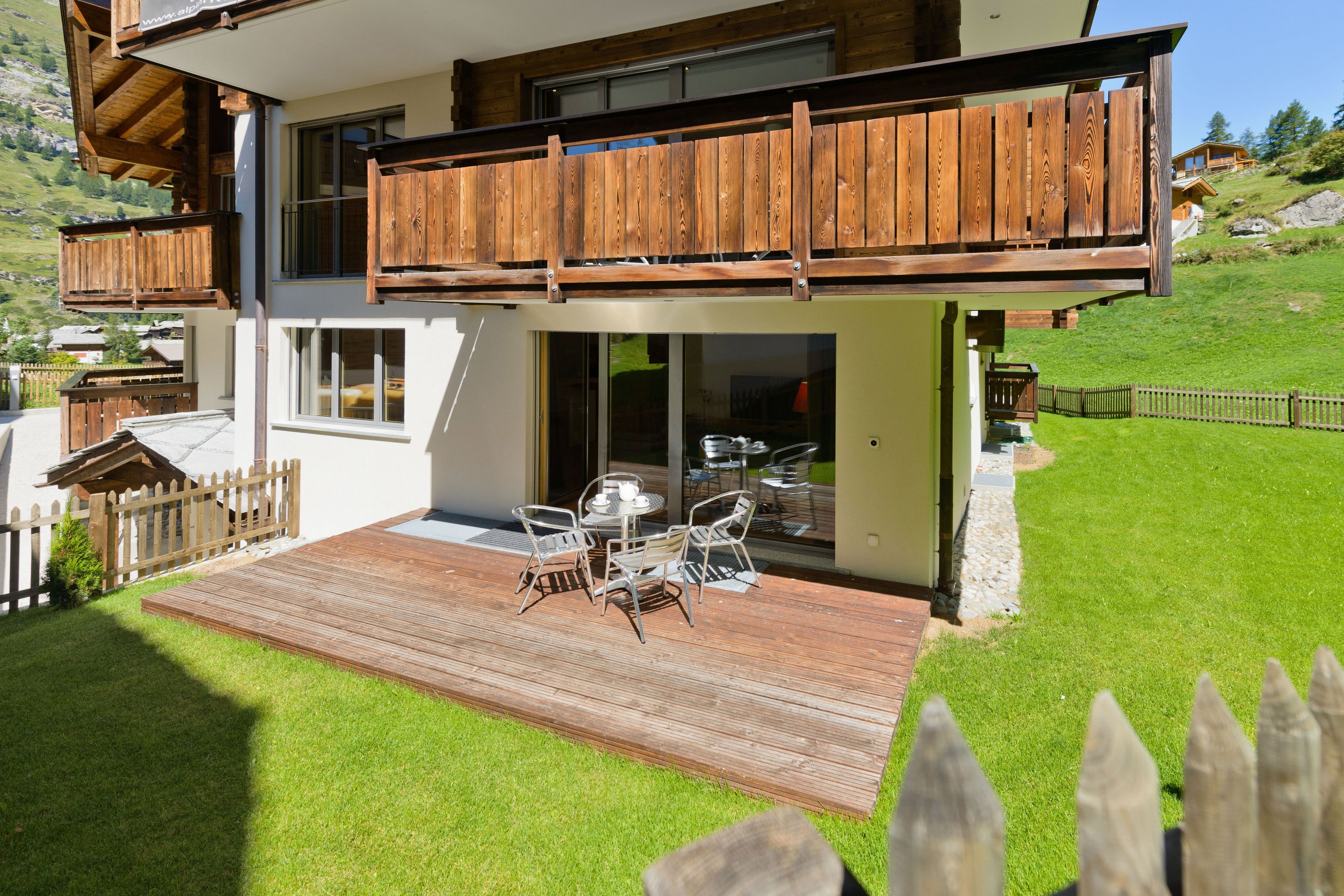 Una casa moderna de dos pisos con una terraza de madera, mobiliario exterior y vistas a una ladera verde.