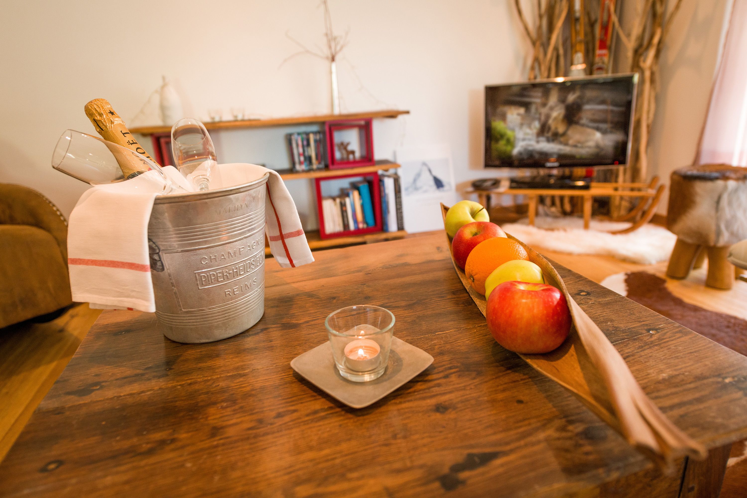 Una mesa de madera con un cubo de champán, una vela y una cuchara de madera sosteniendo manzanas y naranjas.