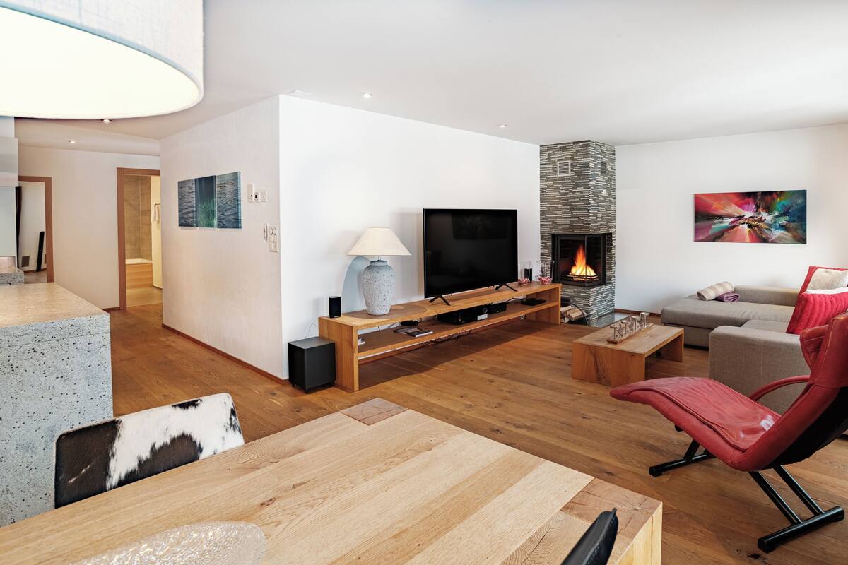 A modern living room with a flat-screen TV, a stone fireplace, a red leather chair, and a grey sofa.