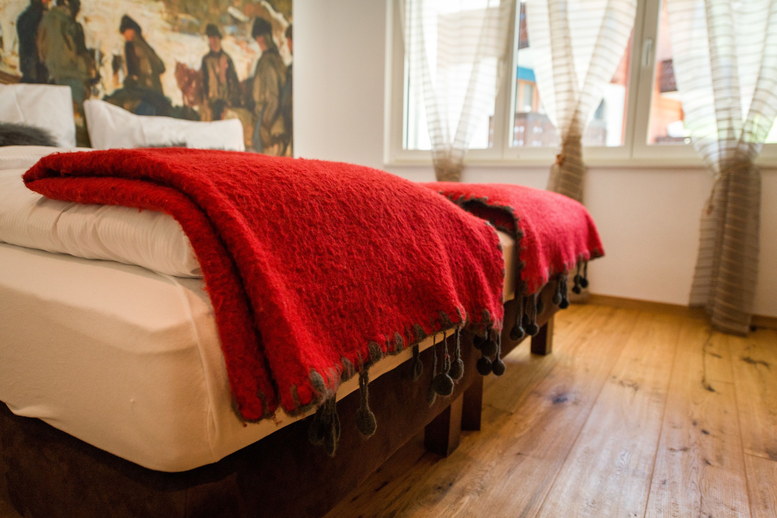 Un lit avec une couverture rouge et des oreillers blancs, un plancher en bois et une fenêtre avec des rideaux.