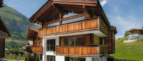 A two-story wooden chalet with a balcony, large windows, and a view of the surrounding mountains.