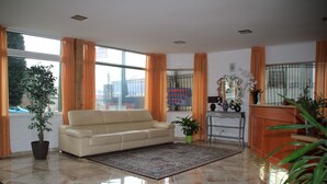 Een ruime woonkamer met een beige bank, een groot raam met oranje gordijnen, een betegelde vloer en een groene plant.