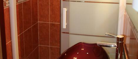 A bathroom with a glass-enclosed shower, a red sink, and a mirror with a light fixture.