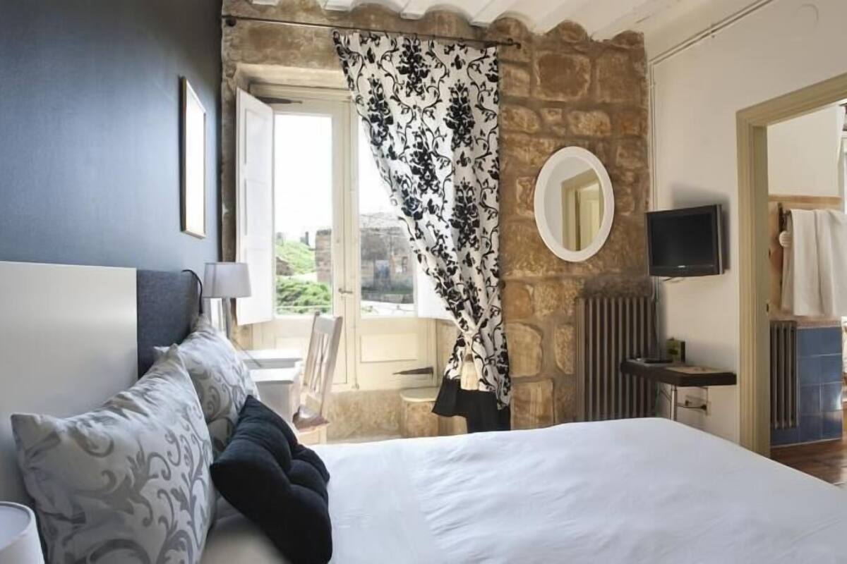 Un dormitorio con cama, televisor, espejo y ventana con cortinas.