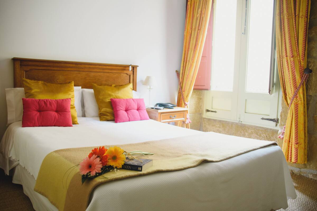 Una cama bien hecha con cabecera de madera, una mesita de noche con lámpara y una ventana con cortinas.