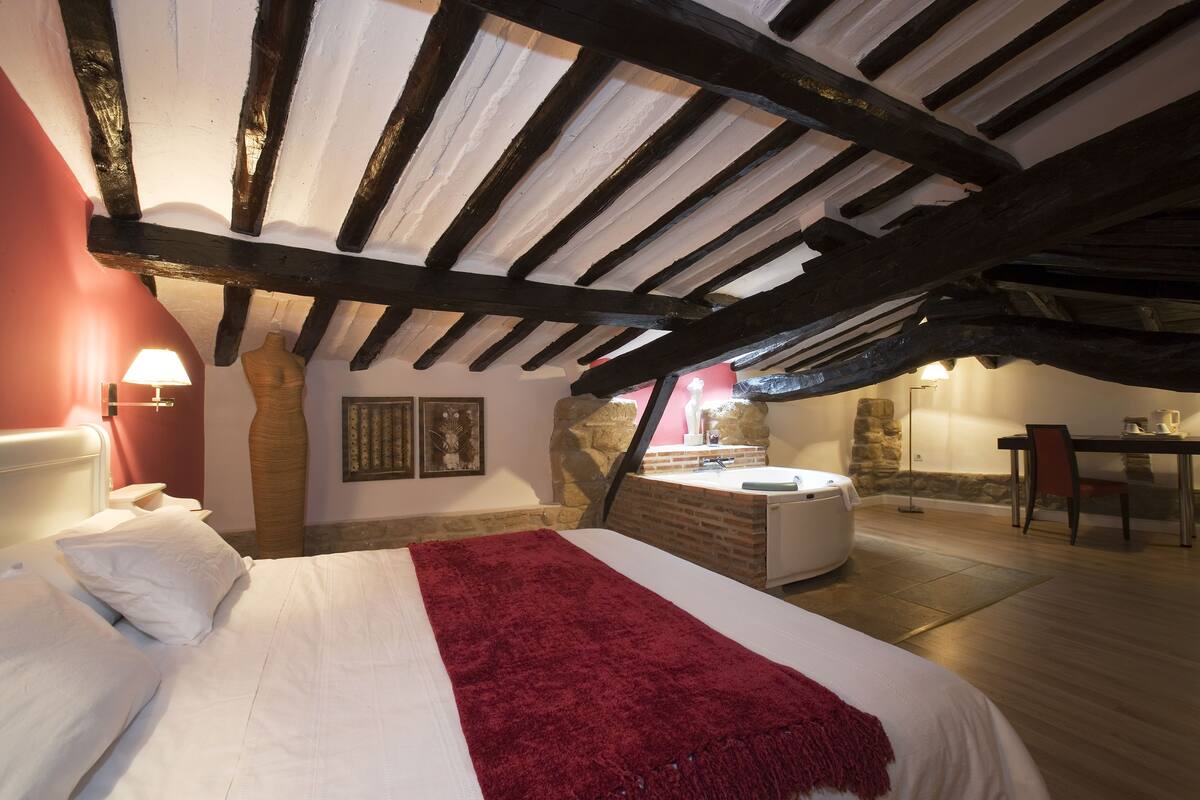 Un dormitorio con una cama grande, una pared de piedra, una chimenea de piedra, una mesa de madera y una silla roja.