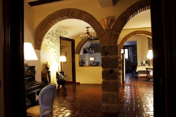 Una habitación con un piano, una silla y una mesa con una lámpara. Se observan dos arcos en el espacio.