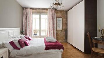 A bedroom with a bed, a wooden floor, a chandelier, a window with curtains, and a desk with a chair.