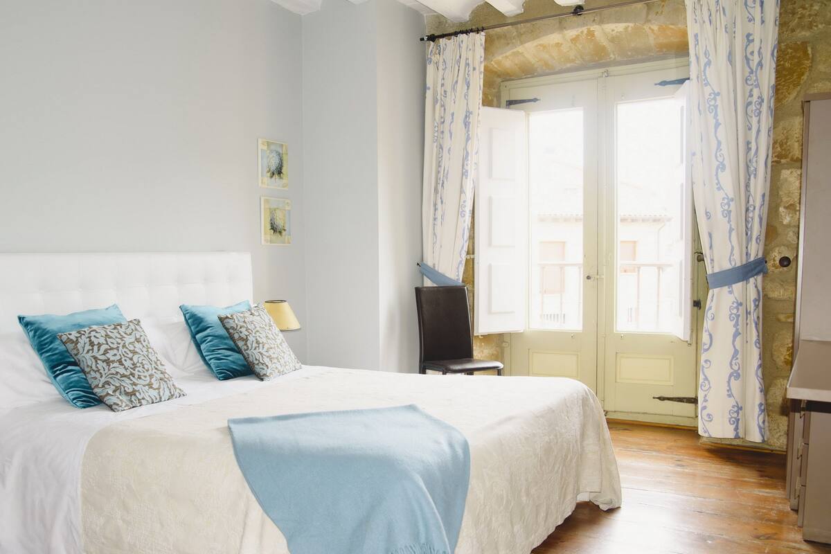 Una cama bien hecha con colcha blanca y azul, dos almohadas y una manta azul. Hay una silla junto a la cama, una lámpara en una mesita de noche y una puerta con cortinas.