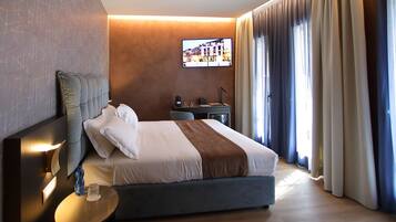 A modern hotel room with a large bed, a bedside table with a lamp, a flat-screen TV mounted on the wall, and a window with curtains.