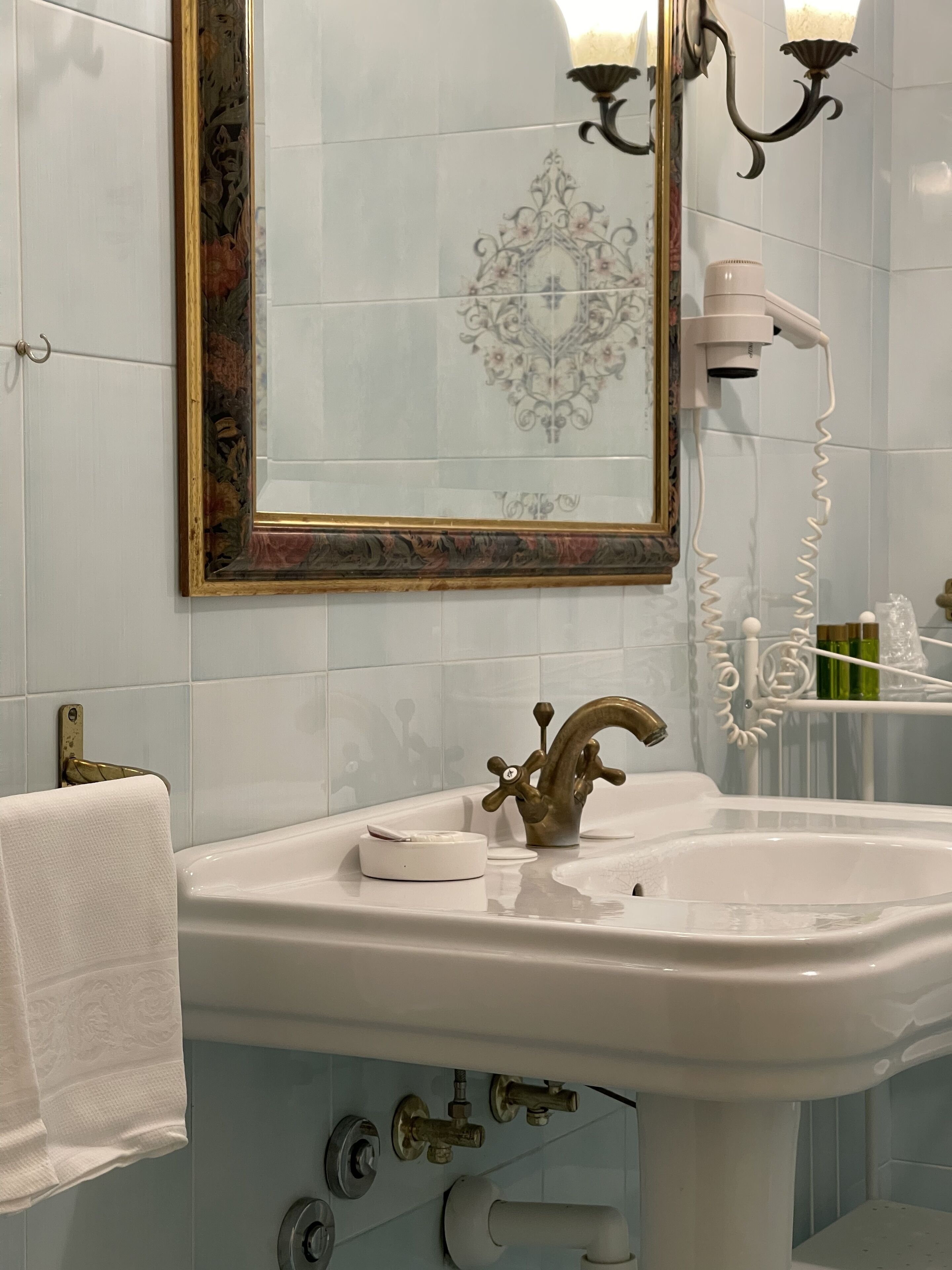 Un bagno con un lavandino bianco, un rubinetto dorato e uno specchio decorativo con cornice dorata.