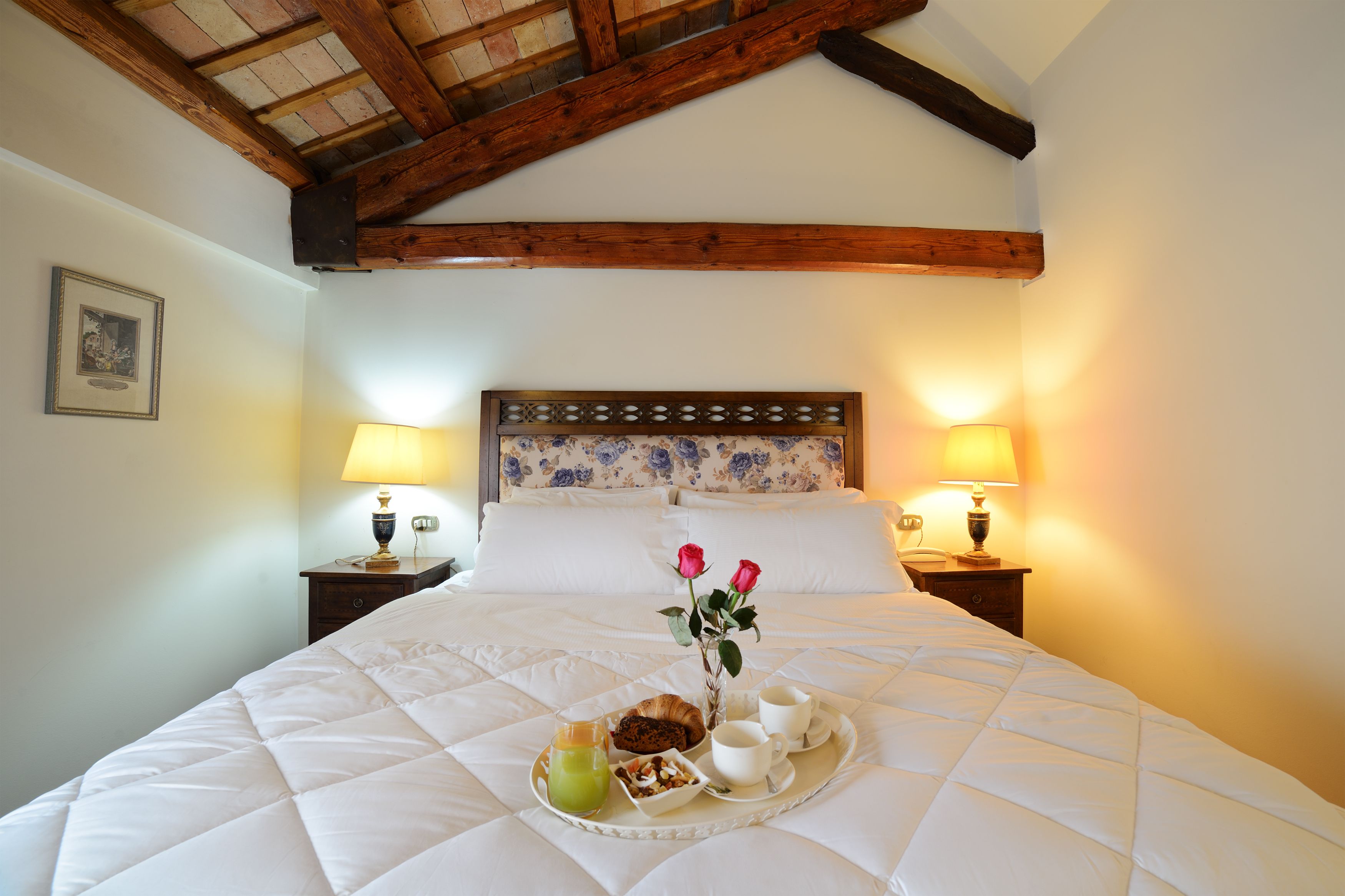 Una cama impecablemente tendida con una bandeja de desayuno, que incluye bollería, café y una bebida, en una habitación con vigas de madera y dos lámparas de noche.