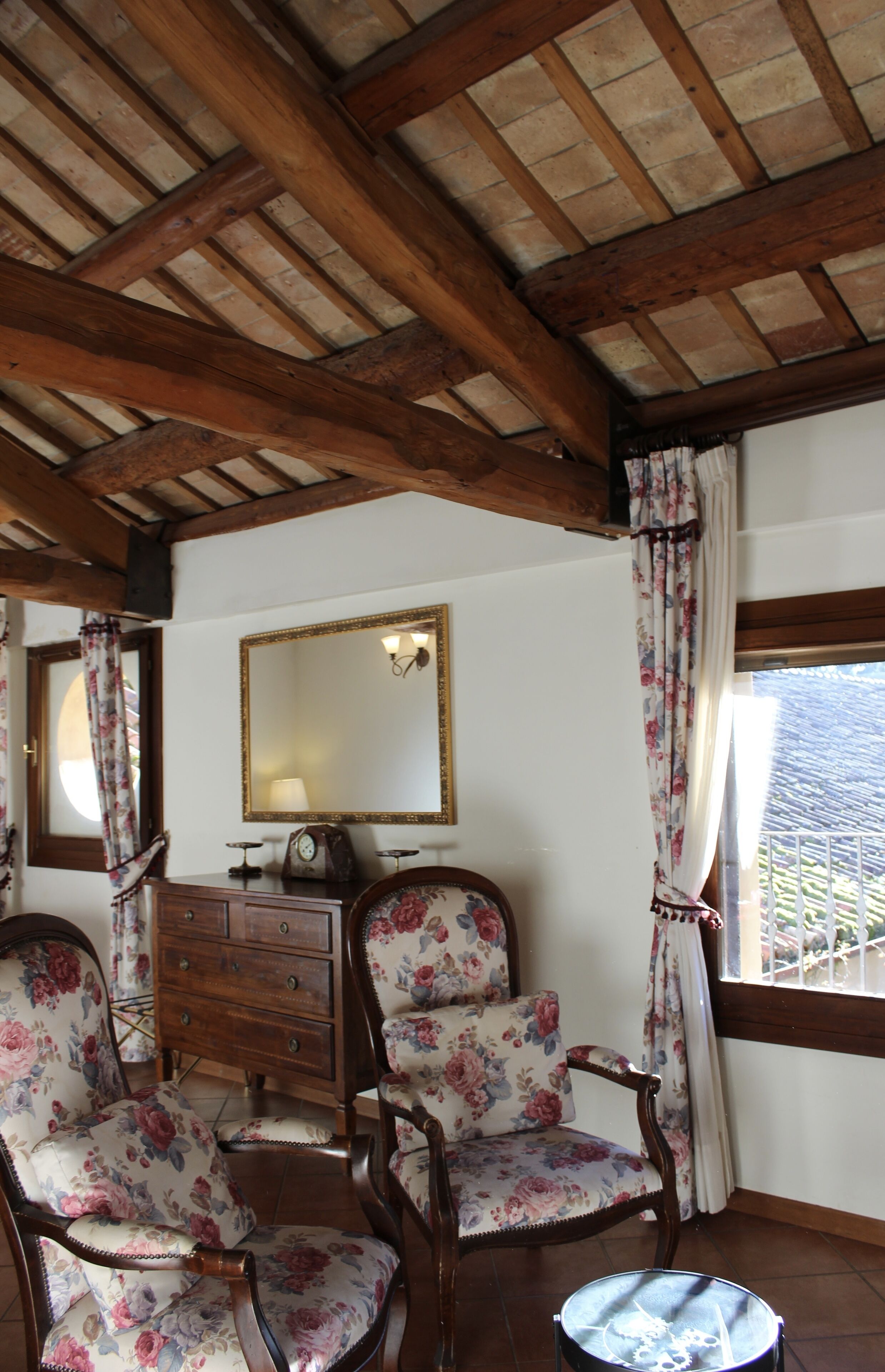Una habitación con vigas de madera en el techo, un aparador con cajones, dos sillones con estampado floral, una mesa redonda y un espejo en la pared.
