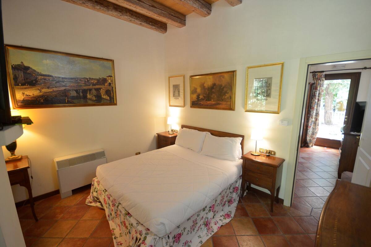 A bedroom with a large bed, two bedside tables, a nightstand, a radiator, and a painting on the wall.