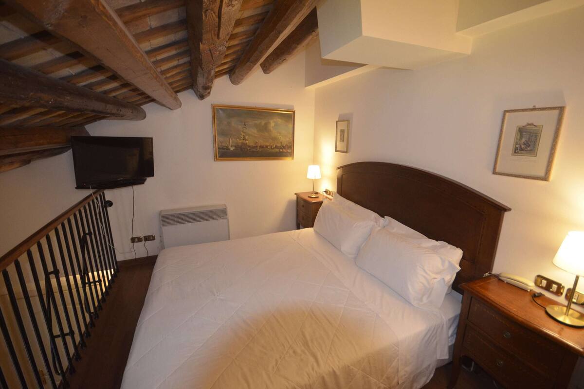 A bedroom with a wooden headboard, a bed with white linens, a bedside lamp, a nightstand, a framed picture on the wall, and a television mounted on the wall.