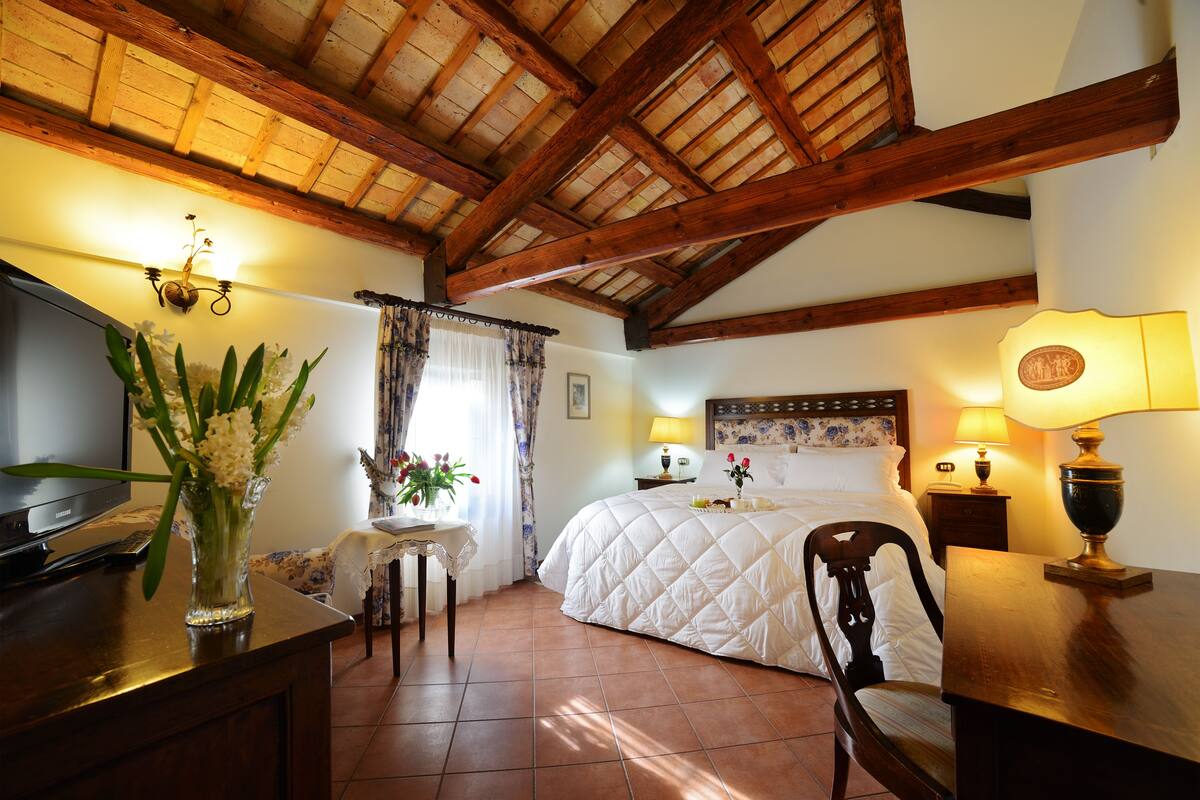 A cozy bedroom with a wooden ceiling, a bed with a quilt, a desk with a lamp, and a vase with flowers.