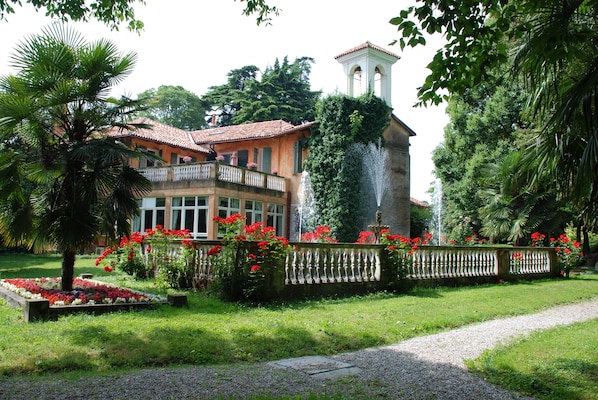 Une grande maison dotée d’un balcon, entourée d’une végétation luxuriante et agrémentée d’une fontaine.