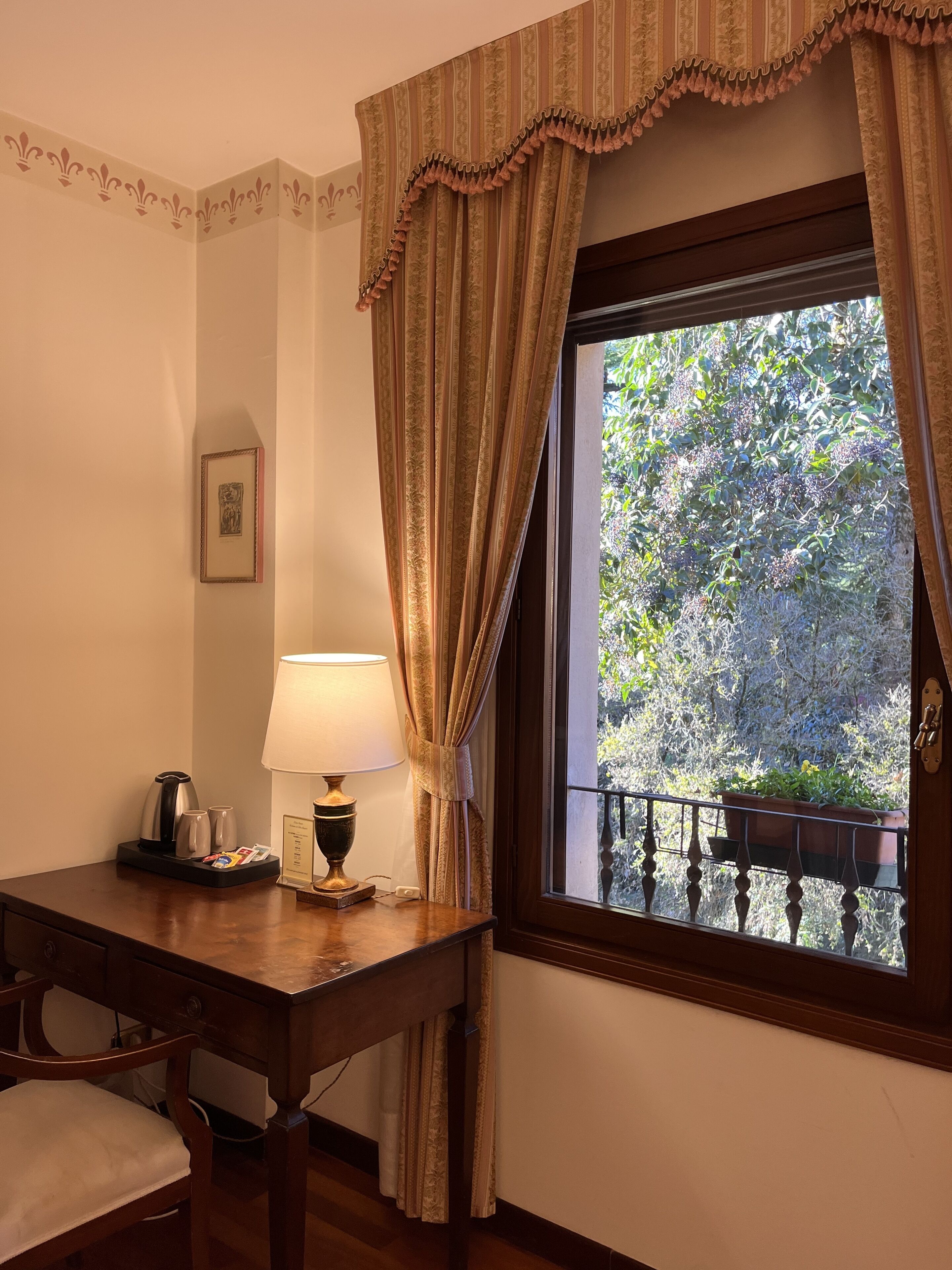 Une chambre d’hôtel avec un bureau en bois, une lampe, un tableau encadré au mur et une fenêtre avec des rideaux.