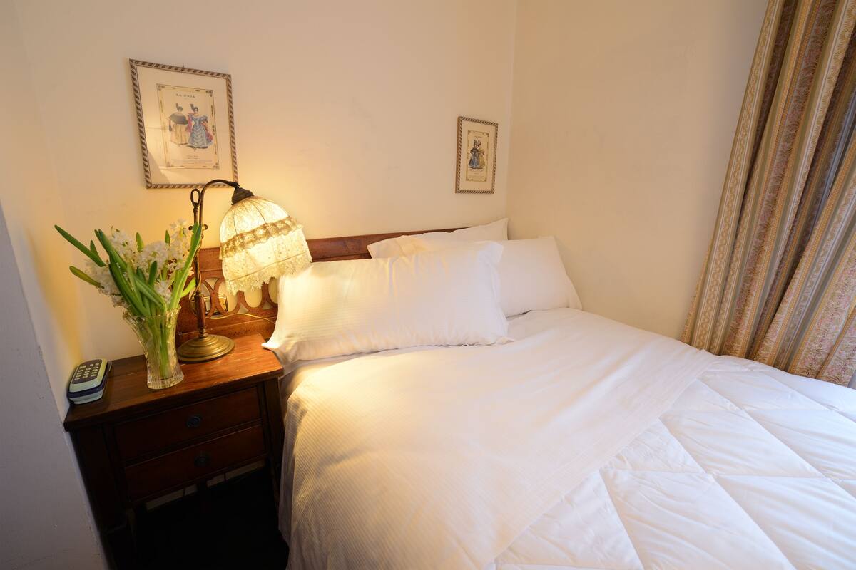 A neatly made bed with white linens, a bedside lamp, a vase with flowers, and two framed pictures on the wall.